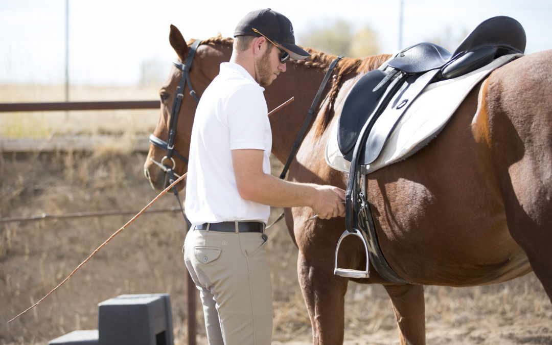 Man in white shirt adjusting horse saddle outdoors.